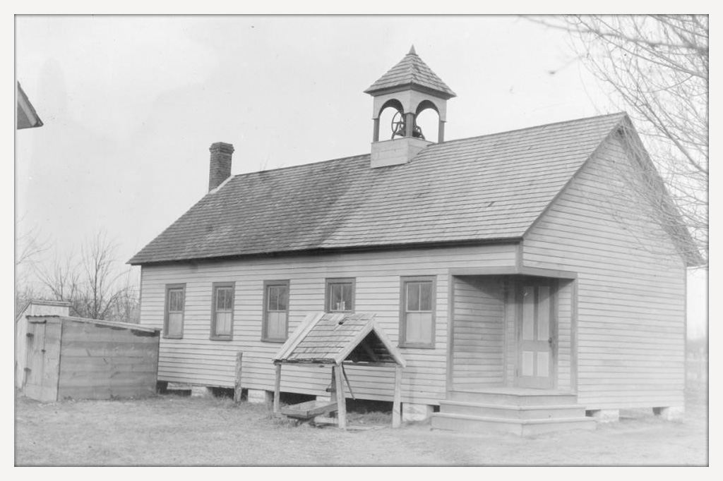 A one-room school with a bell on top and a chicken coup in the yard.