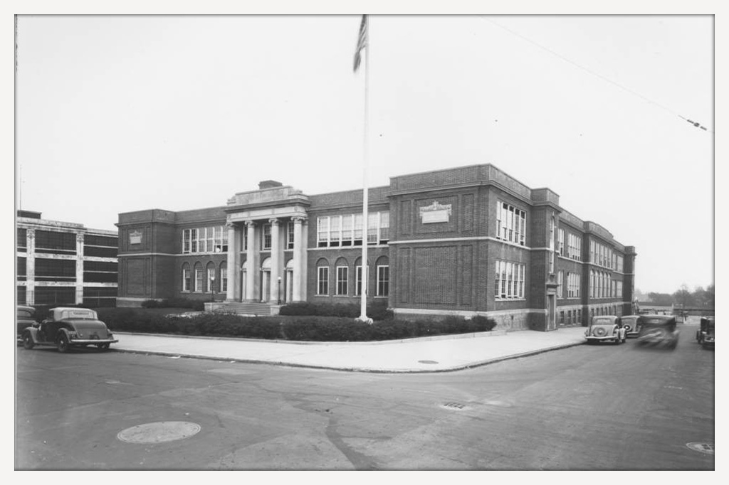 Black-and-white photo of the front of a school building with a flagpole.