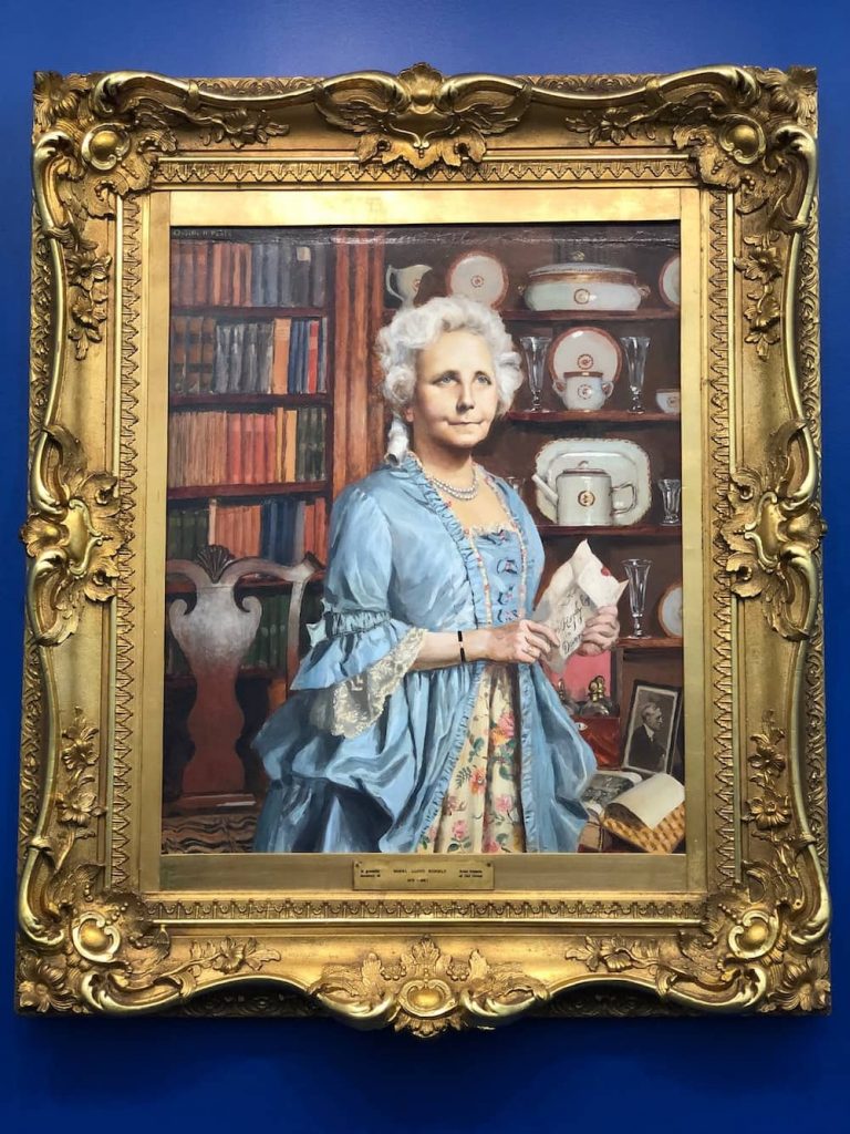 A formal portrait of Mabel Lloyd Ridgely in an elegant blue 18th-century dress, standing with a letter in hand.