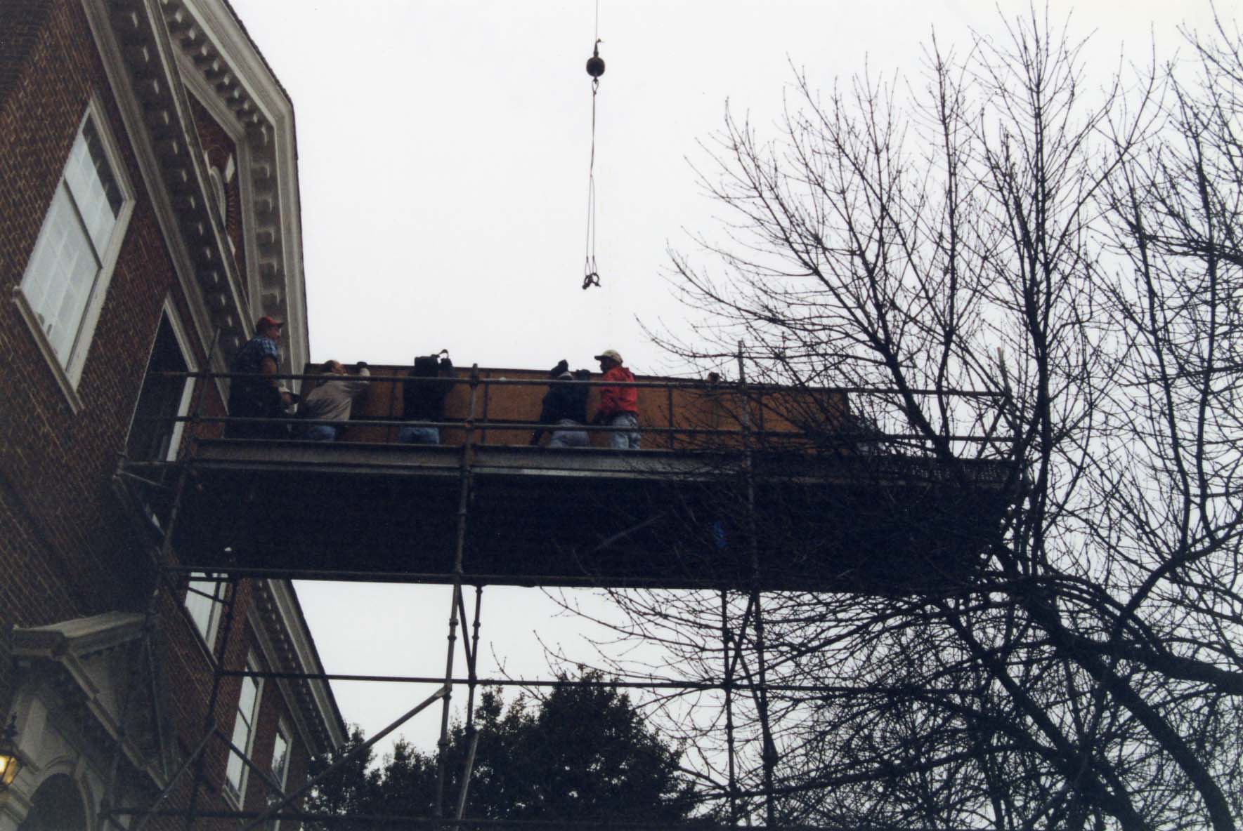 Workers on scaffolding at the Delaware Public Archives are holding a large painting, with a crane hook above and a tree to the right, under an overcast sky.