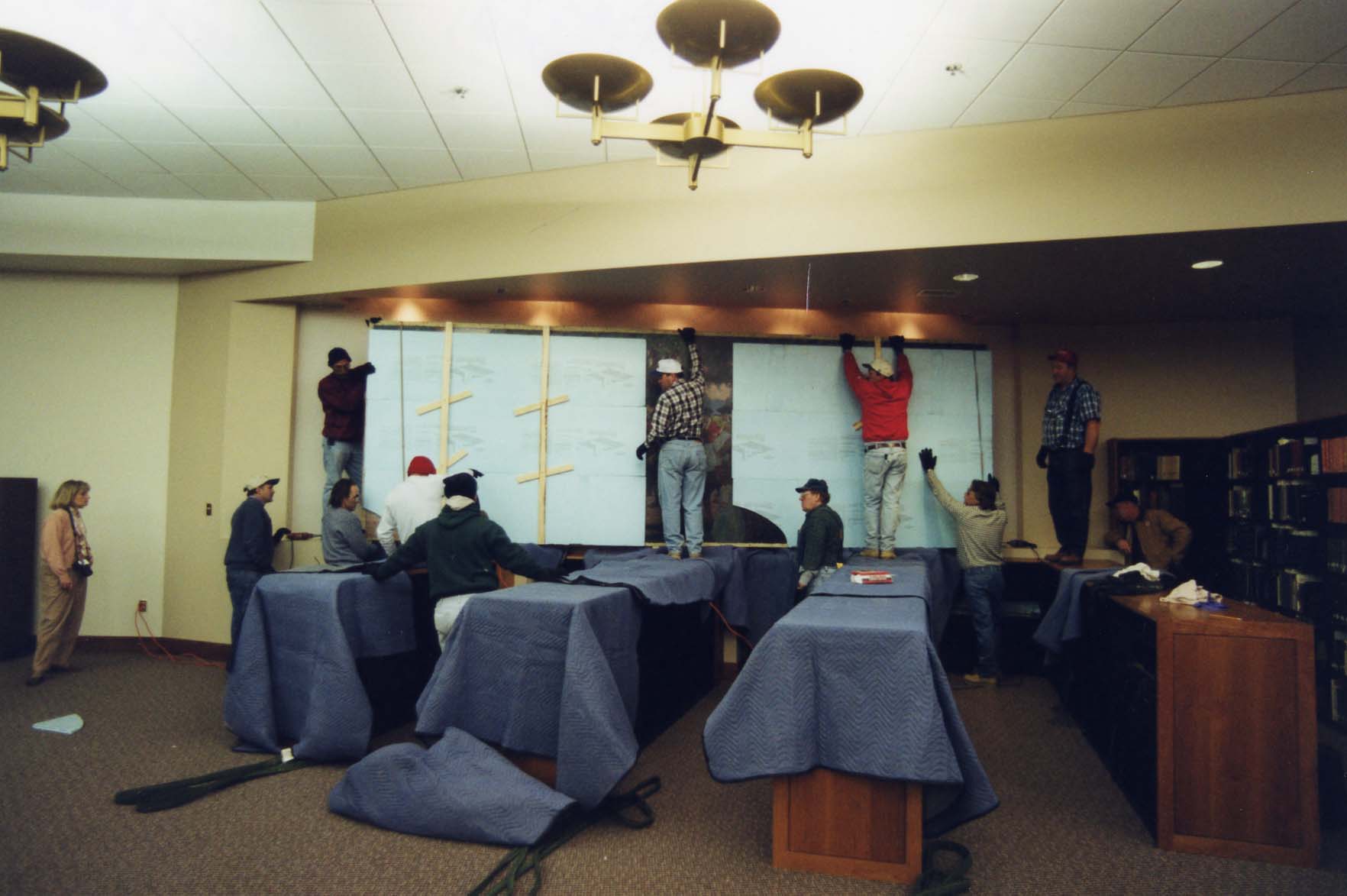 Workers installing Troops Leaving the Green on the wall while standing on covered bookshelves in the Mabel Lloyd Ridgely Research Room.