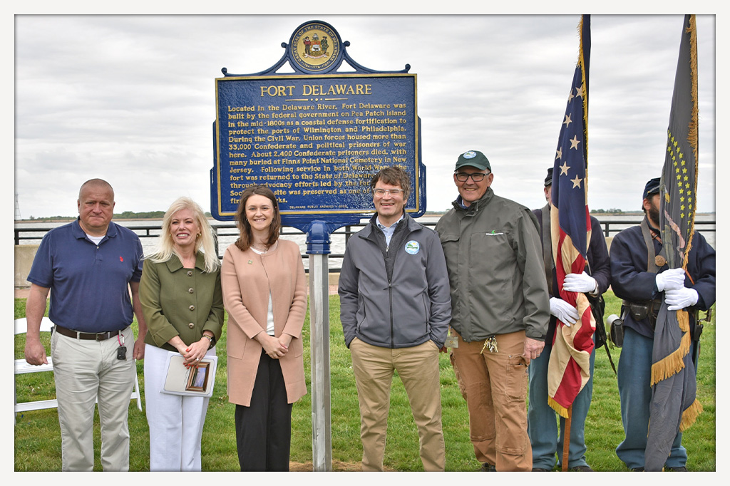 Group of speakers and Delaware State Parks staff standing in front of the newly unveiled Fort Delaware marker with the fort in the background.