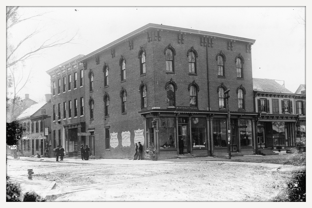 Three-story brick drugstore with large display windows, advertising signs, and several people standing outside, including two children and a baby carriage.