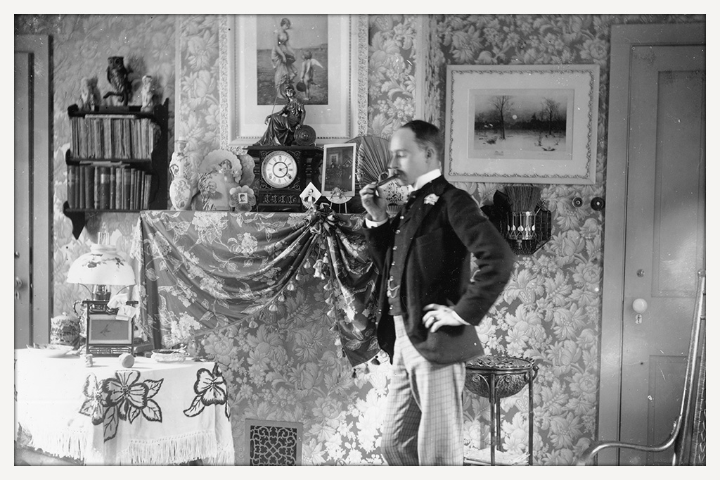 Mustachioed man poses in an ornate parlor, holding a pipe, with framed pictures, books, a rocking chair, and decorated furnishings around him.