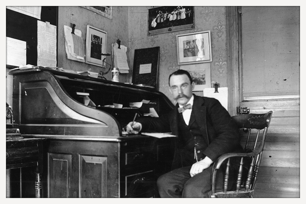 Bow-tied, mustachioed man seated at a roll-top desk, holding a pen, with papers and photographs on the desk and walls behind him.