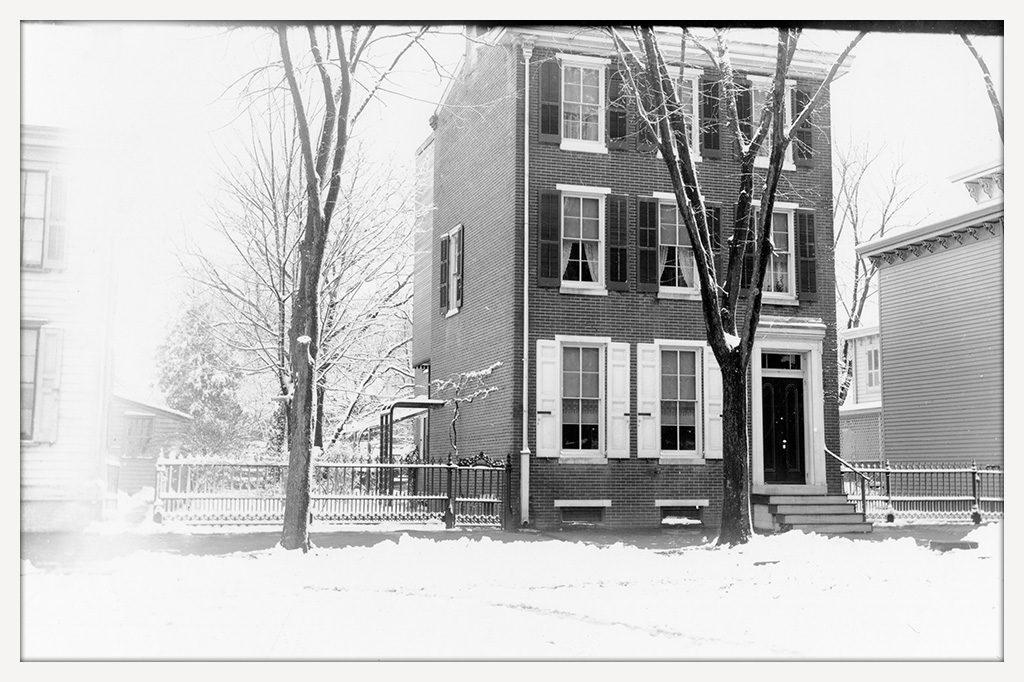 Tall three-story brick house with shuttered windows, flanked by clapboard houses on both sides.