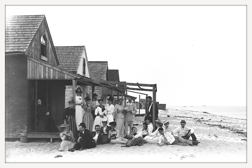 Group of men and women gathered on the sand beside wooden beach cottages, with waves from Delaware Bay reaching the shore nearby.