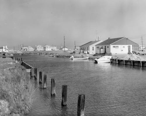 Black-and-white photograph of a mid-century waterfront canal lined with small ranch houses, moored boats, and wooden bulkheads.