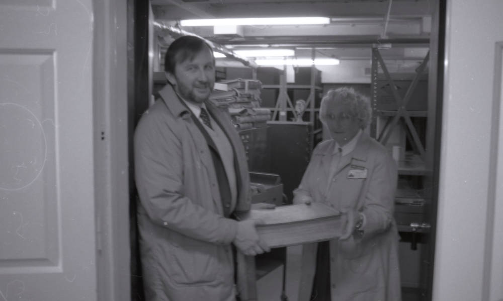 A man and woman maneuver a large book out of a crowded vault.
