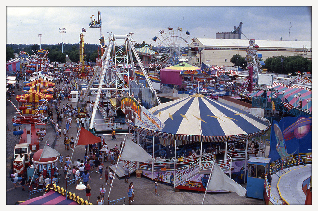 Elevated view of the Delaware State Fair midway, showing crowds, colorful carnival rides, tents, and amusement attractions.