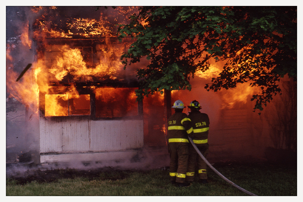 Color photograph of two firefighters directing a hose toward a wooden barn engulfed in flames.