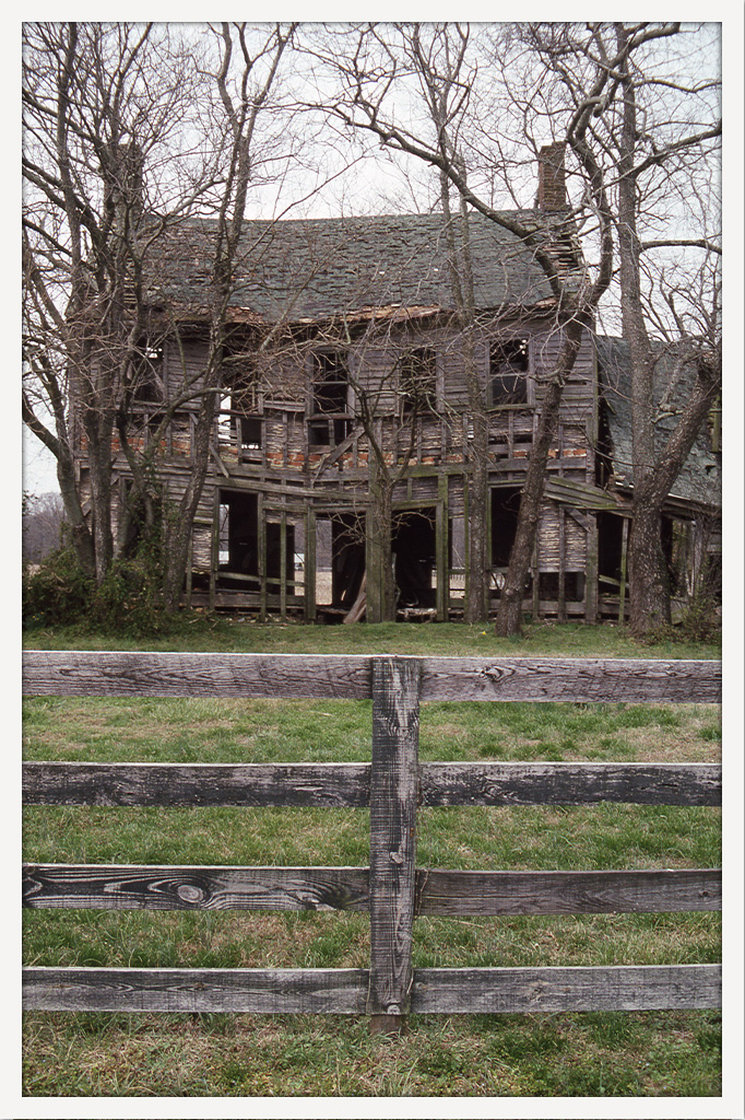 Color photograph of a deteriorating two-story wooden farmhouse with missing windows and siding, seen through leafless trees behind a wooden fence.