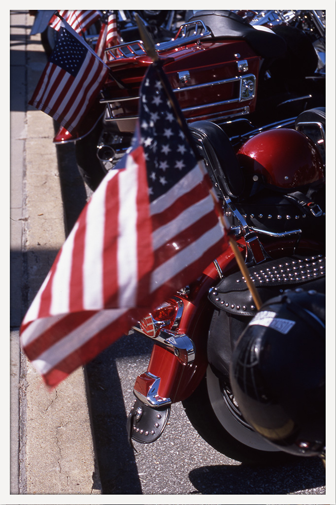 Color photograph of several parked motorcycles decorated with American flags, with one large flag filling much of the foreground.