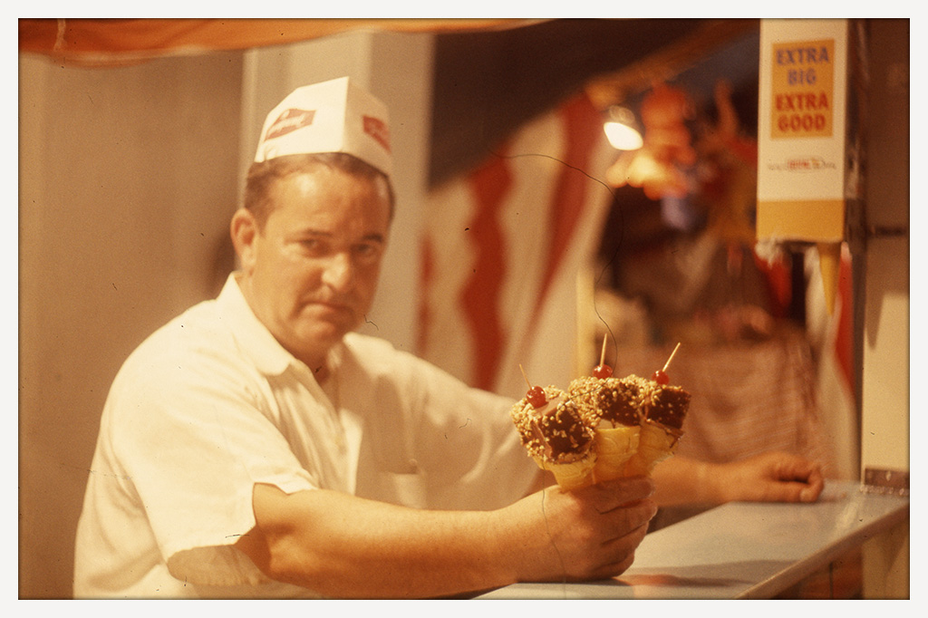 Color photograph of an ice cream vendor in a white shirt and paper hat holding three decorated ice cream cones across a concession counter.
