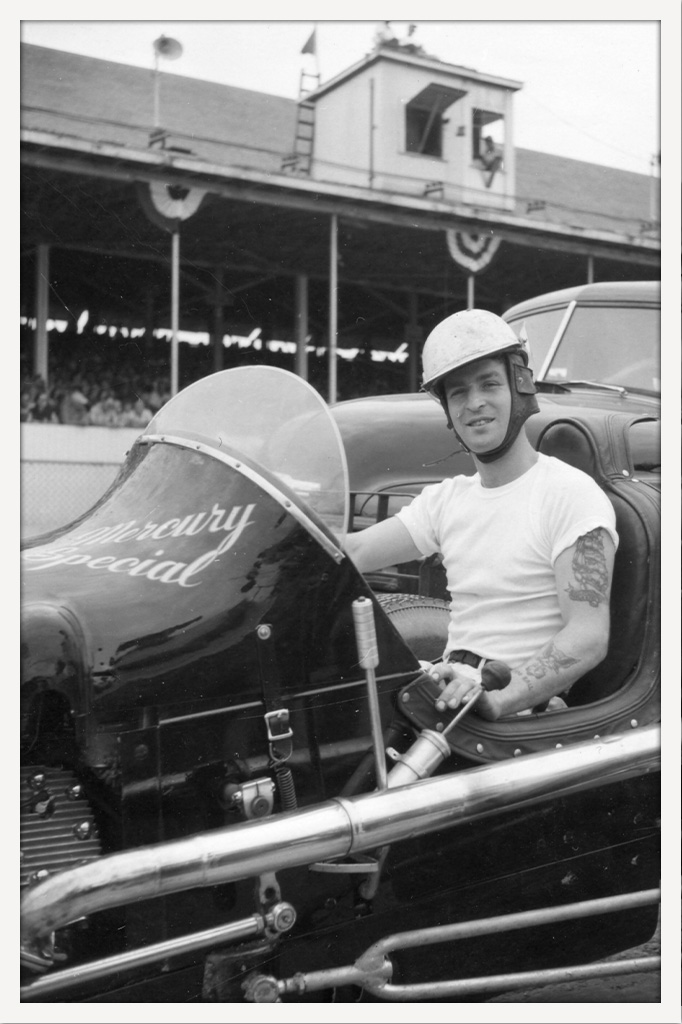 Black-and-white photograph of a helmeted race car driver seated in an open-top car marked “Mercury Special,” with a crowded grandstand behind him.