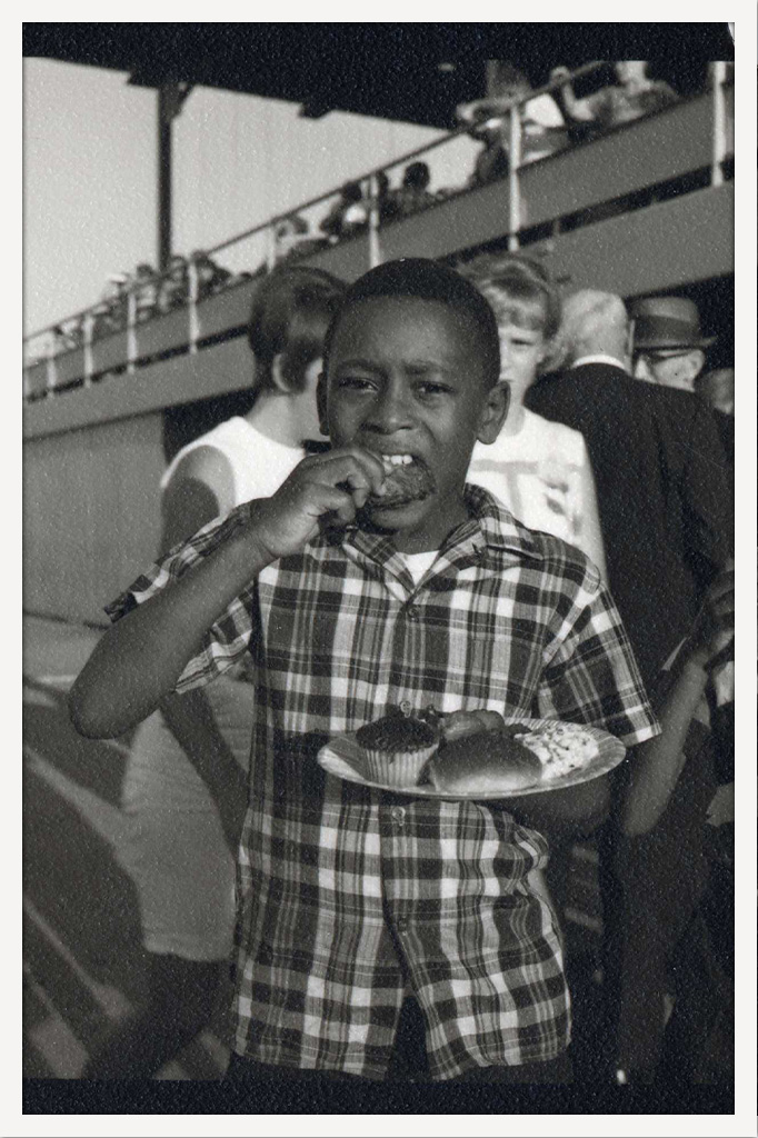 Black-and-white photograph of a young boy in a plaid shirt taking a bite of food while holding a paper plate with a cupcake and other food.