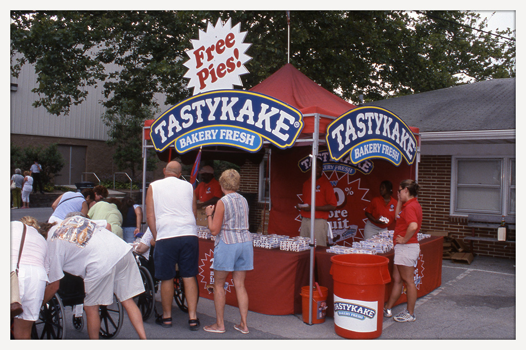 Color photograph of fairgoers gathered around a red Tastykake concession booth with large blue-and-white logos and a sign reading “Free Pies!”