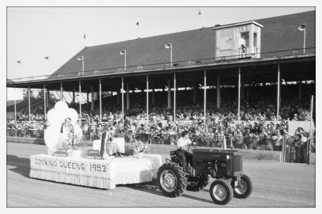 Black-and-white photograph of a tractor pulling a “Cooking Queens 1953” parade float carrying two crowned women past a crowded fair grandstand.
