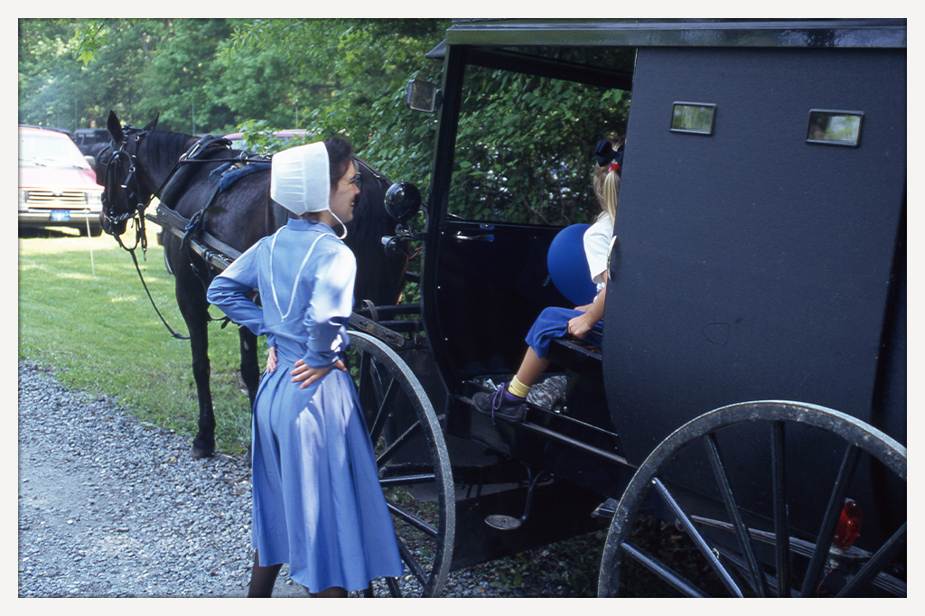 Color photograph of a young girl holding a blue balloon inside a black horse-drawn buggy while a woman in a blue dress and white bonnet stands beside it.