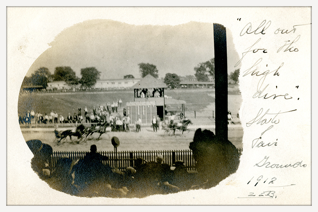 Black-and-white postcard showing harness racers passing a trackside viewing stand as spectators watch from the infield and shaded grandstand.