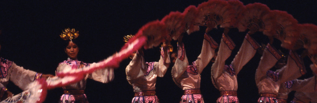 Performers in pink gowns holding fans in an arched formation during a fan dance.