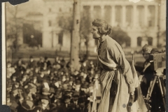 Suffragist Lucy Branham in an Occoquan Prison dress in 1919. Branham was speaking at an outdoor meeting during the National Woman’s Party “Prison Special” tour. She is above a large crowd, wearing a prison dress and suffrage sash, with a suffrage flag and camera on a tripod right behind her.